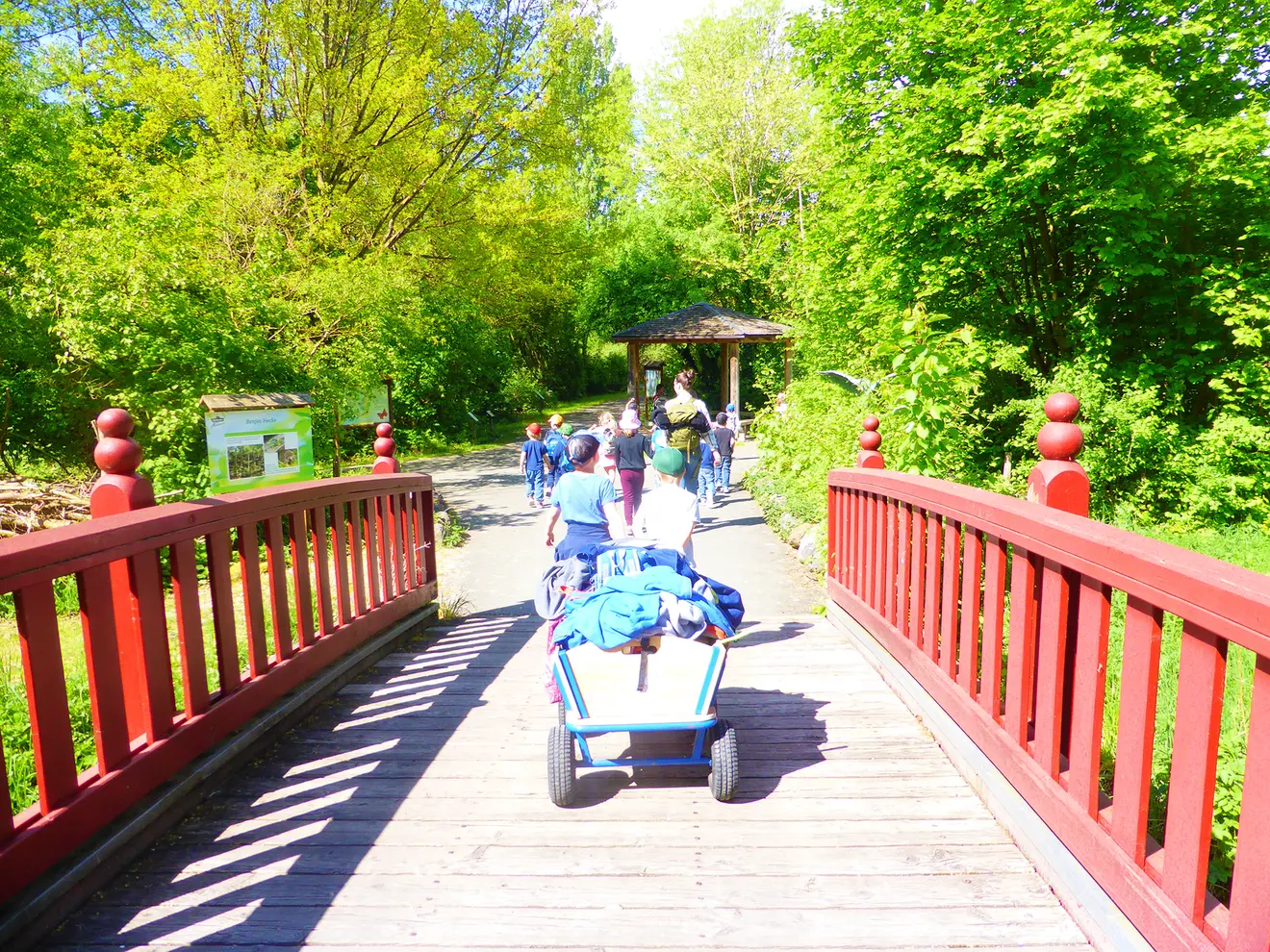 Gruppe von Kindern und Erwachsenen auf einer Holzbrücke im Grünen, im Hintergrund ein Pavillon.