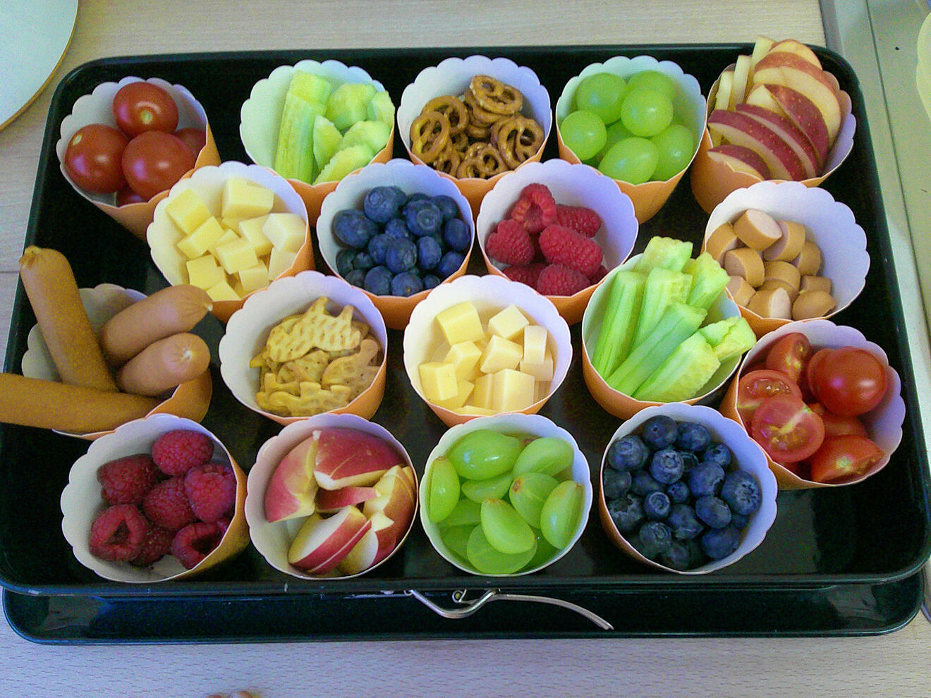 Vielseitige Snack-Platte mit Obst, Gemüse, Käse, Brezeln und Würstchen in bunten Förmchen.