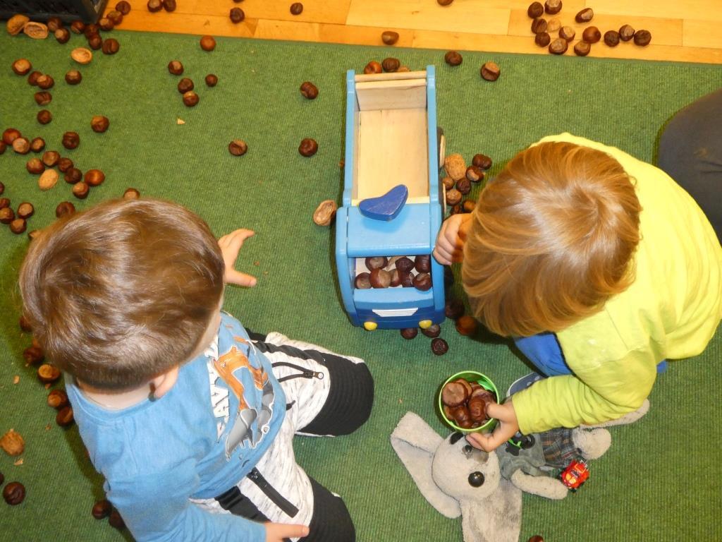 Zwei Kinder spielen mit Kastanien und einem blauen Spielzeugauto auf einem grünen Teppich.