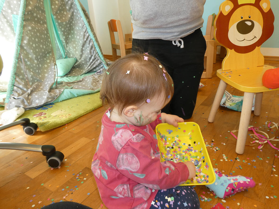 Kleines Kind sitzt auf dem Boden und spielt mit bunten Konfetti in einer gelben Schüssel.