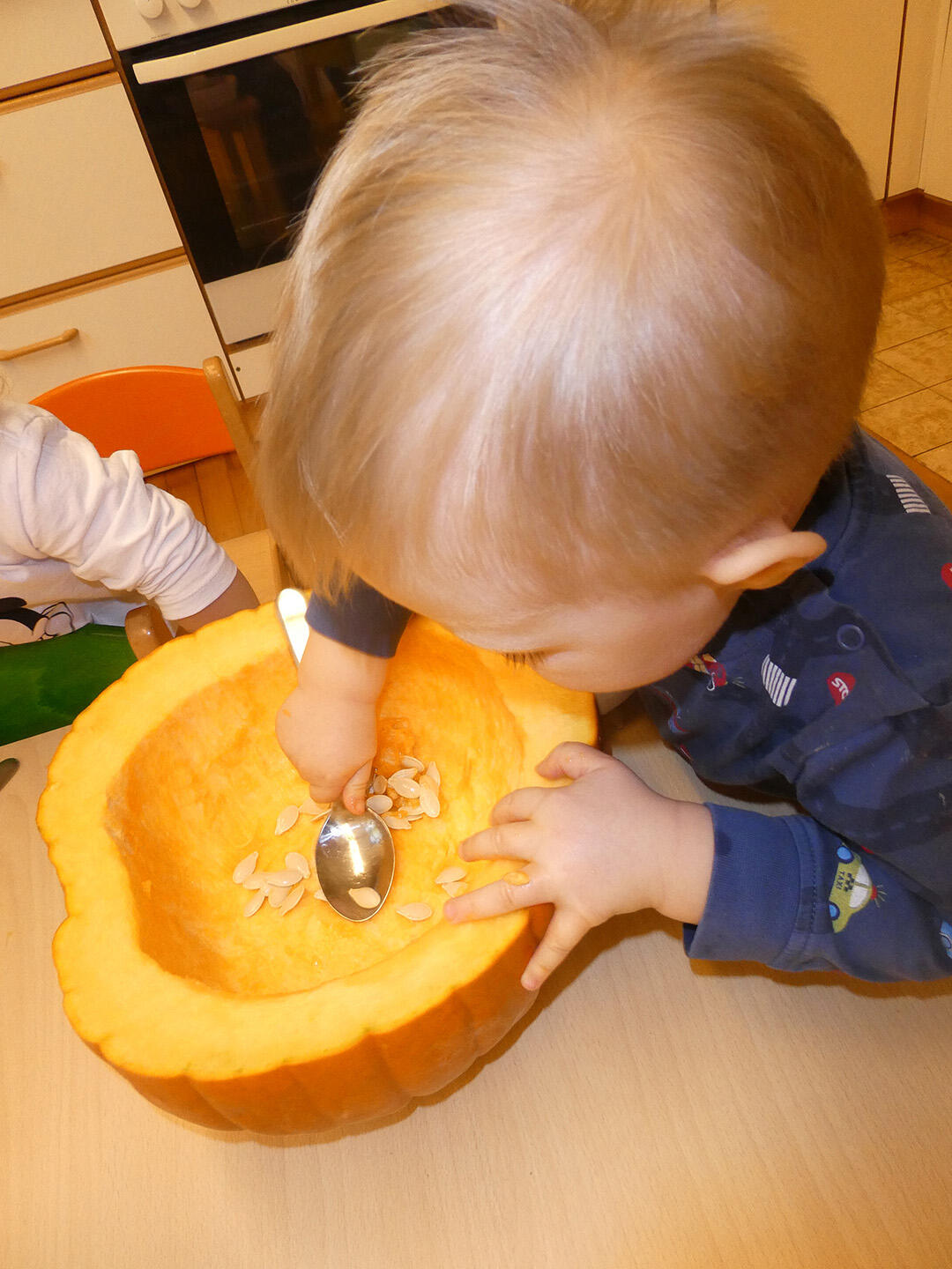 Ein Kind schöpft mit einem Löffel Kürbiskerne aus einem großen, orangefarbenen Kürbis.