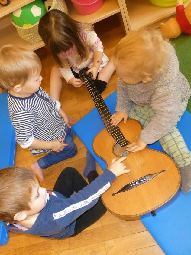 Vier Kinder spielen gemeinsam mit einer Gitarre auf dem Boden in einem Raum.