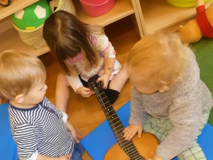 Vier Kinder spielen gemeinsam mit einer Gitarre auf dem Boden in einem Raum.
