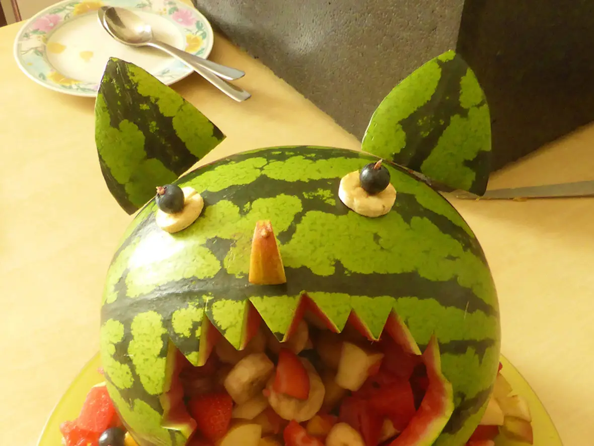 Eine kreativ gestaltete Melone mit Fruchtfüllung als Monster, umgeben von buntem Obst.