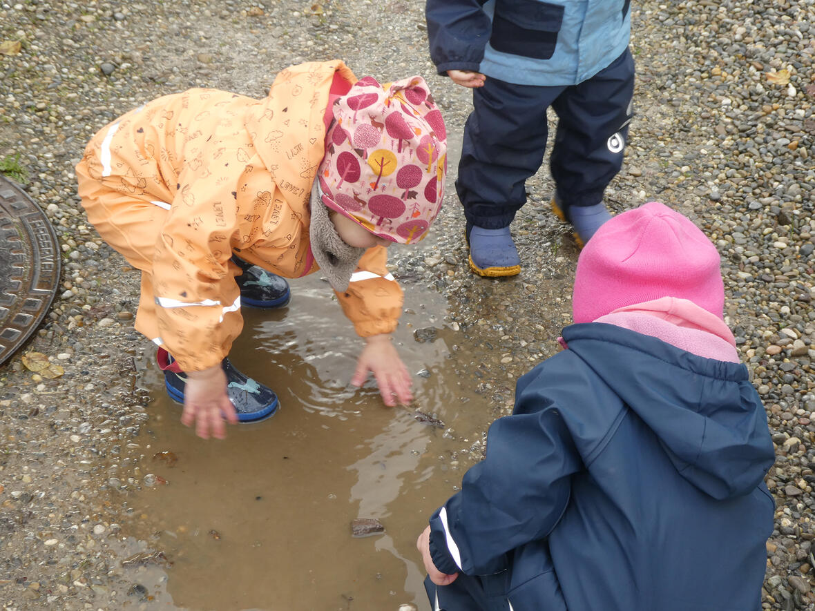 Kinder spielen in einer Pfütze, während eine von ihnen mit den Händen im Wasser plantscht.