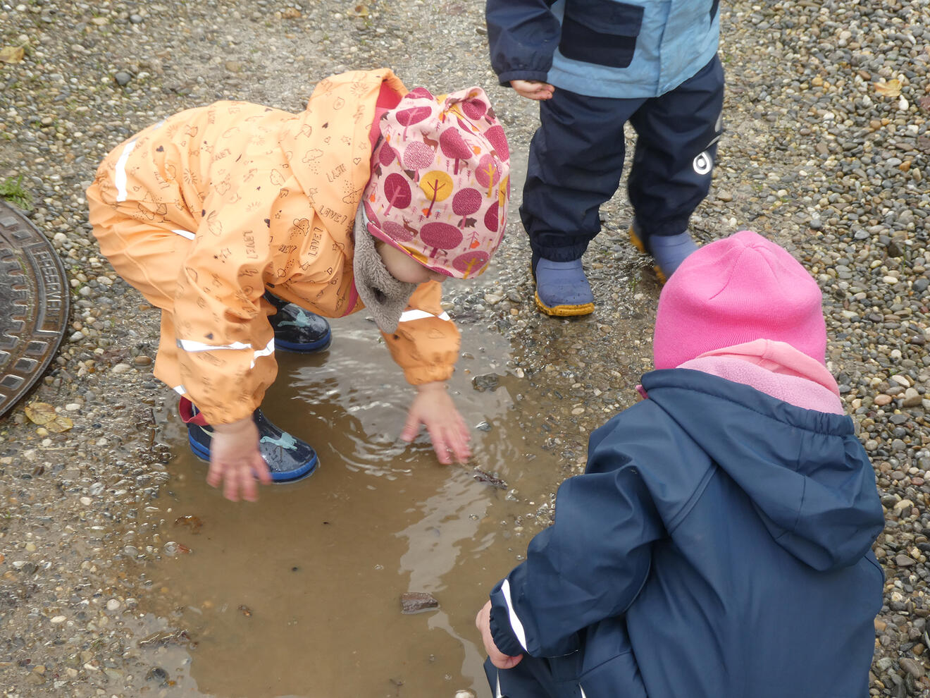 Kinder spielen in einer Pfütze, während eine von ihnen mit den Händen im Wasser plantscht.