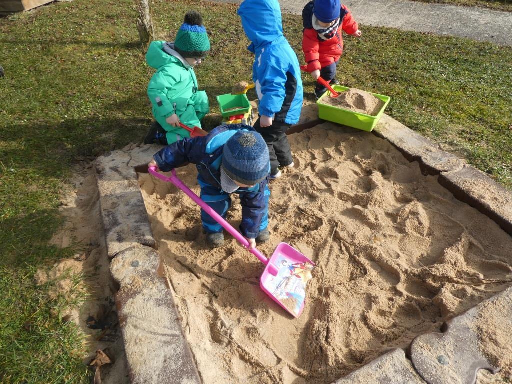 Vier Kinder spielen im Sandkasten und benutzen Schaufeln, um Sand zu transportieren.