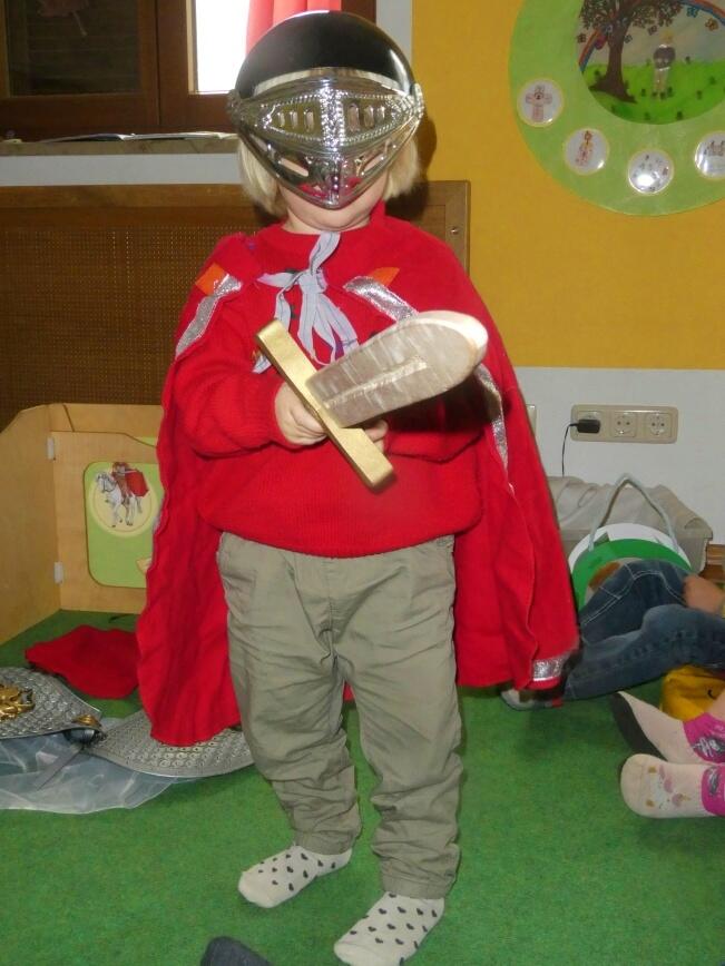 Kleines Kind mit Helm und rotem Umhang, hält ein Holzschwert in einem Spielzimmer.