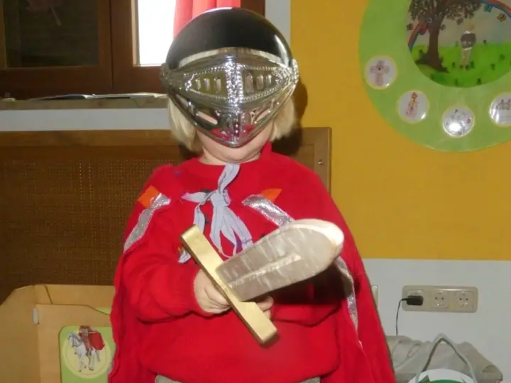 Kleines Kind mit Helm und rotem Umhang, hält ein Holzschwert in einem Spielzimmer.
