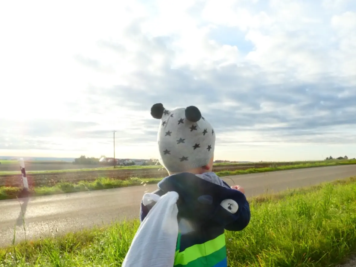 Kind mit einem Kapuzenpulli und einem Tuch, steht an einer Landstraße und schaut in die Ferne.