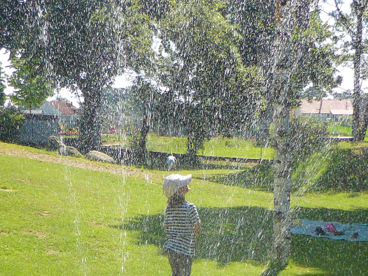 Ein Kind mit einem weißen Hut spielt im Sprühnebel eines Wasserspenders im Park.