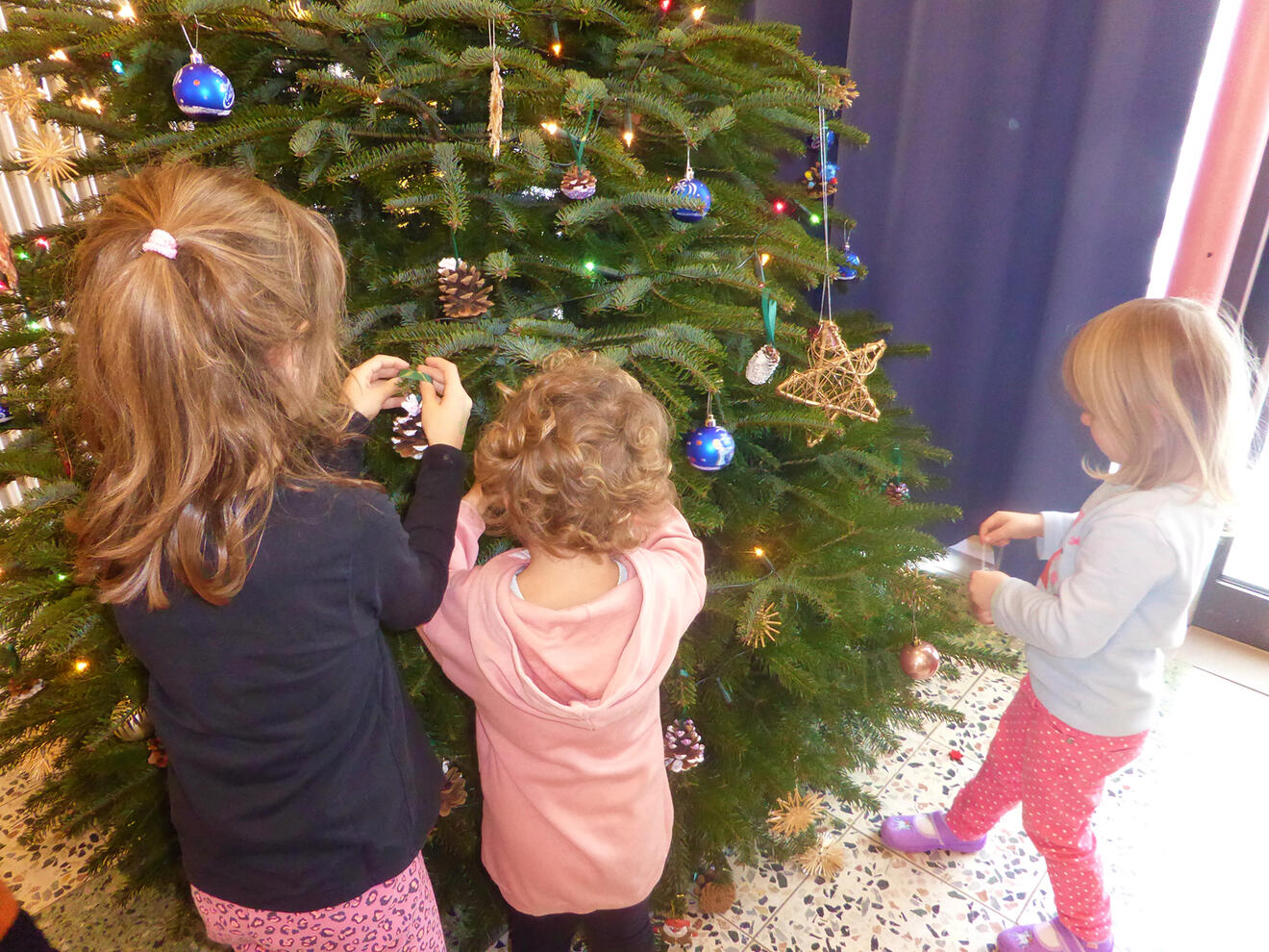 Drei Kinder schmücken einen Weihnachtsbaum mit buntem Schmuck und Naturmaterialien.