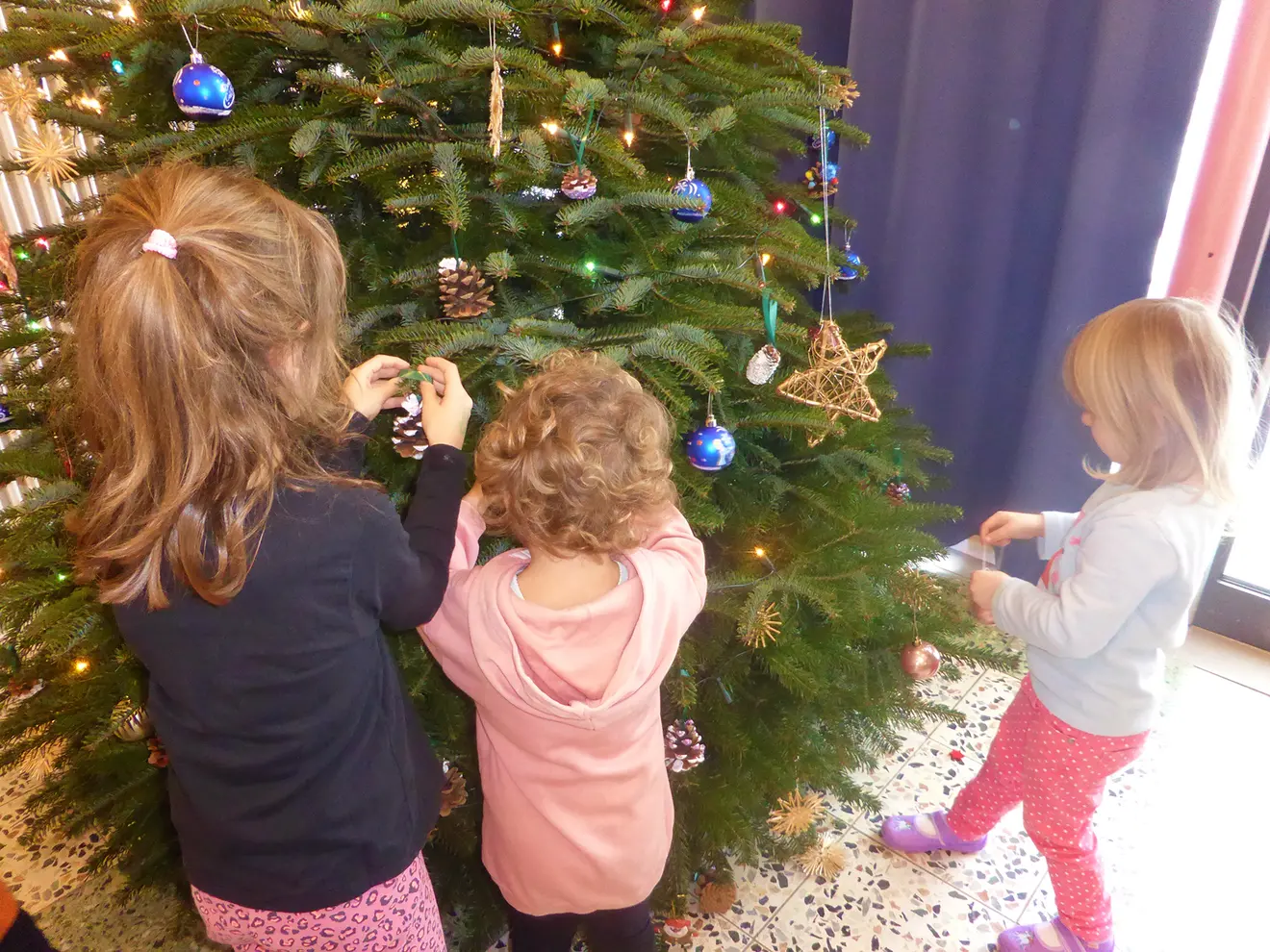 Drei Kinder schmücken einen Weihnachtsbaum mit buntem Schmuck und Naturmaterialien.