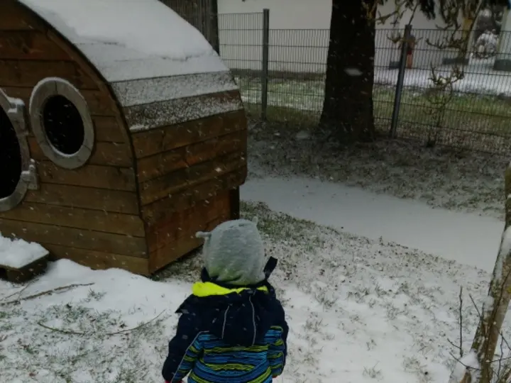 Ein kleiner Junge steht im Schnee neben einem roten Spielzeugauto und einer Holzhütte.
