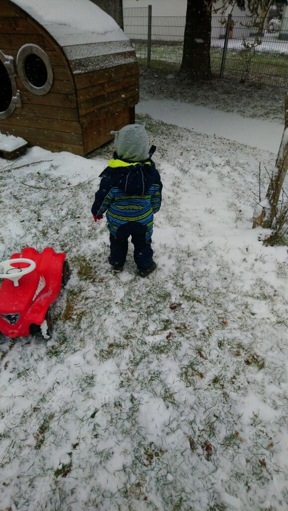 Ein kleiner Junge steht im Schnee neben einem roten Spielzeugauto und einer Holzhütte.
